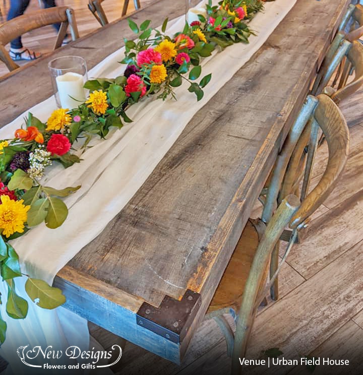 a colourful garland of exotic and wild flowers in deep purples and oranges, atop spiky greenery runs along a soft white chiffon table runner, along a long wooden table.